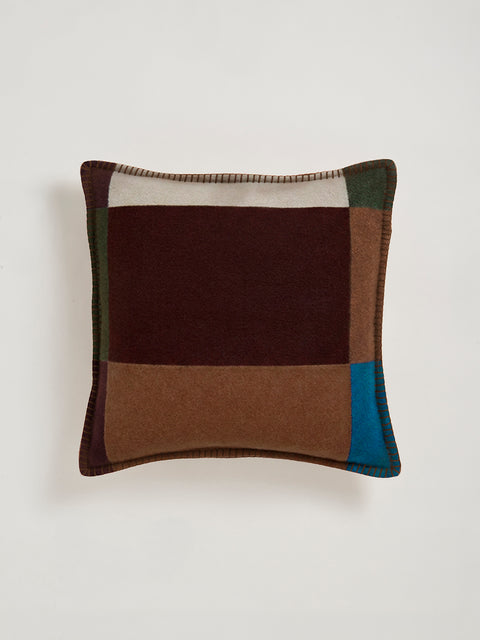 A square decorative throw pillow with a patchwork design in burgundy, brown, blue, and green, featuring a stitched border, shown against a neutral light gray background.