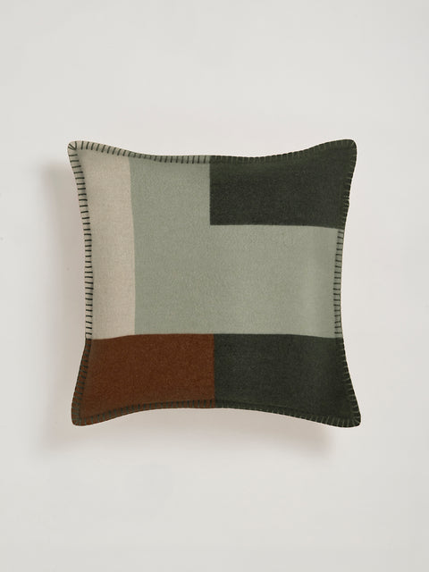 A square throw pillow with a patchwork design in sage green, olive green, brown and beige, featuring a dark green stitched border, shown against a neutral light gray background.