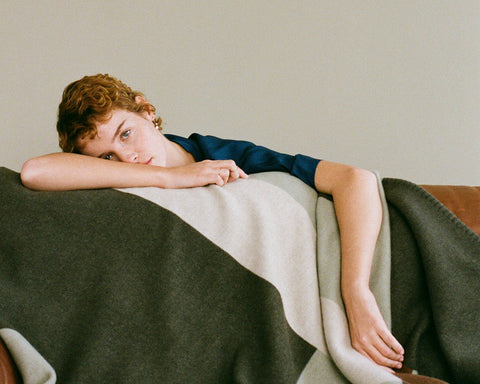 A woman with short curly red hair lies draped over a brown leather couch covered with a large knit blanket. The blanket features a color-blocked design in dark olive green, light sage green and cream.