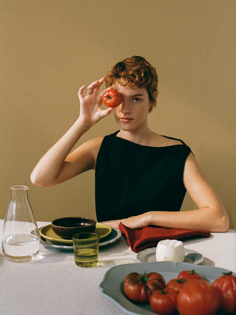 A model with short curly red hair wearing a black sleeveless top sits at a table holding a red tomato to her eye. The table is styled with plates, a glass carafe, a green glass, and a bowl of fresh tomatoes.