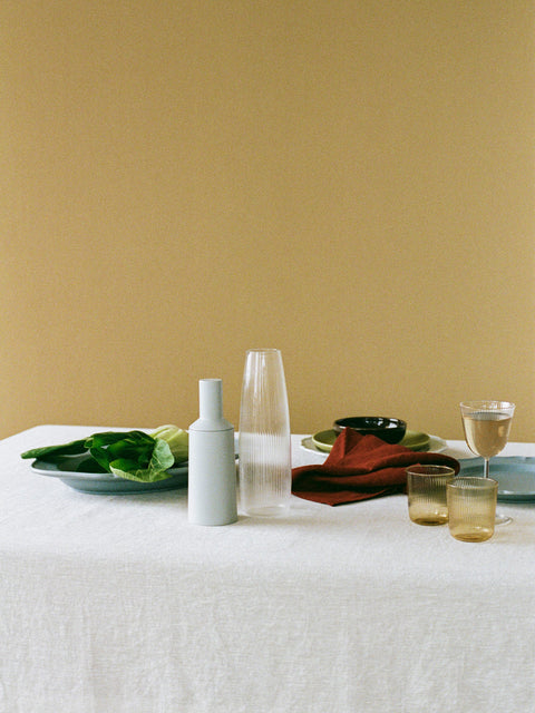 A styled tabletop scene with a tan linen tablecloth set in front of a dark yellow wall. The table has a pale blue plate of green leaves, a ribbed glass carafe, a pale blue pepper mill, a folded rust-red napkin, small amber-tinted glasses, and a wine glass and various plates and bowls.