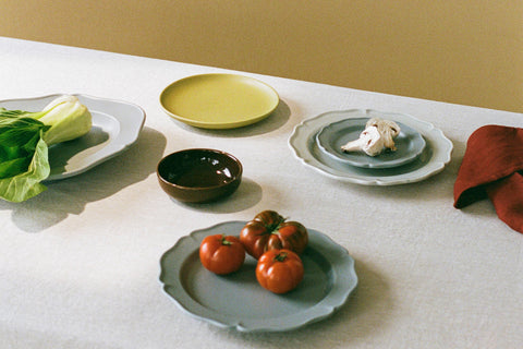 A table with beige linen tablecloth in front of mustard wall, set with various blue, green and brown ceramic plates, one with tomatoes, another with bok choy and third with mushrooms.