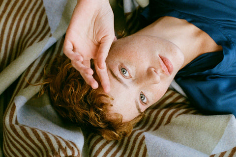 A close-up lifestyle shot of a person lying down, looking directly at the camera with their hand near their face, wearing a blue top and resting on a striped beige and brown blanket.
