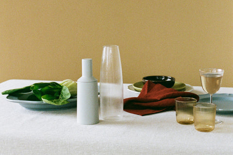 A styled tabletop scene with a tan linen tablecloth set in front of a dark yellow wall. The table has a pale blue plate of green leaves, a ribbed glass carafe, a pale blue pepper mill, a folded rust-red napkin, small amber-tinted glasses, and a wine glass and various plates and bowls. 